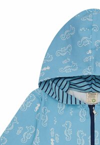 Felpa con zip di colore azzurro chiaro con motivi di cavallucci marini bianchi. La parte interna del cappuccio ha una fodera a righe blu navy e bianche. Materiale morbido in cotone.