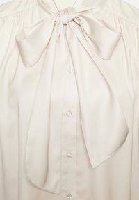 Blusa color crema con un grande fiocco al colletto, dalla texture satinata e morbida, con chiusura frontale a pulsanti, caratterizzata da cinque bottoni.