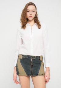 Λευκό πουκάμισο με μακριά μανίκια συνδυασμένο με μίνι φούστα από τζιν με αντιθετικά μπλε και μπεζ πάνελ, φερμουάρ μπροστά και μεταλλικές λεπτομέρειες.