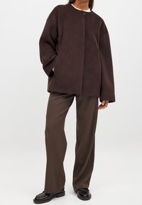 Brauner Fleece-Pullover mit Rundhalsausschnitt, lockerem Schnitt und langen Ärmeln, kombiniert mit braunen weit geschnittenen Hosen und schwarzen Schuhen.