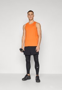 Oranje sporttanktop met mesh-details, gecombineerd met zwarte shorts en leggings, houdt een waterfles vast, draagt zwarte sneakers.