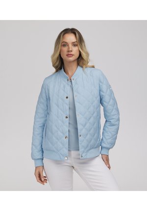 Kobieta w jasnoniebieskiej pikowanej kurtce zapinanej na zatrzaski, stojąca z jedną ręką na biodrze na tle jednolitego szarego tła.