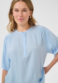 Camicia azzurro chiaro a maniche corte realizzata in tessuto morbido, con scollatura rotonda e patta con bottoni; presenta dettagli arricciati sulle spalle.