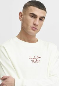 Biały sweatshirt z długimi rękawami i okrągłym dekoltem, z haftowanym napisem "La dolce Vita COTE D'AZUR" w kolorze brązowym.