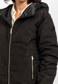 Giubbotto imbottito nero con un interno in tessuto lucido, zip dorate e tasche laterali. Presenta un cappuccio e un logo sulla manica sinistra.