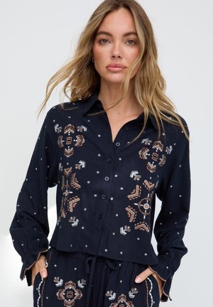 Blonde Frau mit offenem Haar, die eine marineblaue Knopfleistenbluse und passende Hosen mit brauner und weißer geometrischer Stickerei trägt.