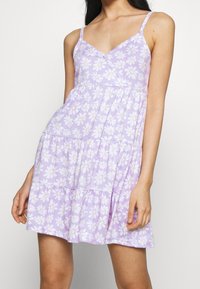 Fioletowa sukienka w kwiaty z białymi stokrotkami, z dekoltem w kształcie V, regulowanymi ramiączkami i warstwowym fasonem spódnicy. Wykonana z miękkiej bawełny.