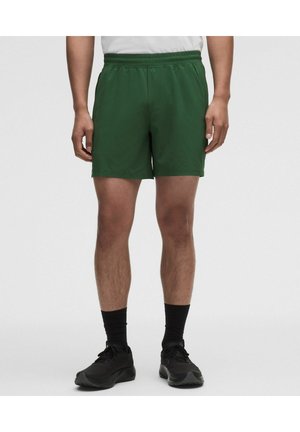 Hombre con pantalones cortos deportivos verdes, calcetines negros y zapatillas negras sobre un fondo liso claro.