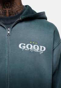 Felpa con zip verde scuro realizzata in tessuto morbido, con la scritta bianca "GOOD For Nothing" e tre stelle gialle sopra di essa.