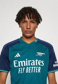 Maglietta da calcio in teal e blu navy con colletto, caratterizzata da testo bianco "Emirates Fly Better", logo Adidas ed emblema del club sul davanti.