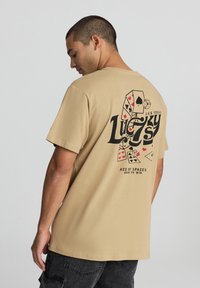 T-shirt in cotone beige con stampa grafica nera di dadi e carte da gioco sul retro, con il testo "Lucky" e "Las Vegas."