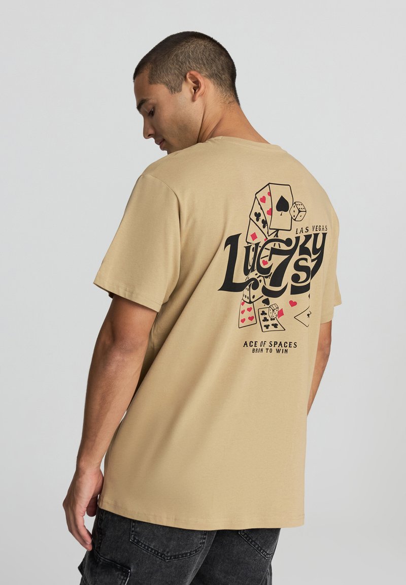 T-shirt in cotone beige con stampa grafica nera di dadi e carte da gioco sul retro, con il testo "Lucky" e "Las Vegas."