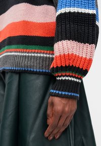 Main reposant sur le côté portant un pull tricoté rayé multicolore et une jupe en cuir vert foncé.