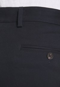 Detailní záběr na černé kalhoty na míru s knoflíkovou zadní kapsou a poutkem na opasek na pasu.