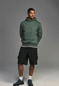 Sudadera verde con capucha y bolsillo delantero, combinada con pantalones cortos de cargo negros. Usada con zapatillas deportivas negras y calcetines blancos hasta el tobillo.