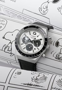 Montre en acier inoxydable argentée avec cadran blanc, accents noirs, échelle tachymètre, bracelet en cuir noir et graphique Snoopy sur le cadran.