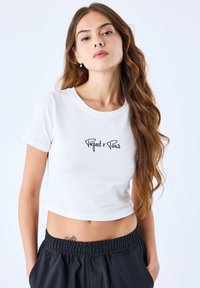 T-shirt blanc court avec un col rond et un design de texte noir "Project x Paris" sur la poitrine ; porté avec un pantalon noir à taille élastique.