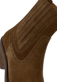 Brown suede ankle boot with vertical stitching, perforated details, and a stacked heel. Smooth texture and curved design.