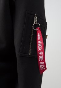 Schwarzer Jackenärmel mit einer vertikalen Reißverschlusstasche, einem silbernen Reißverschlussanhänger, der an einem roten Schlüsselring mit der Aufschrift "REMOVE BEFORE FLIGHT" befestigt ist.