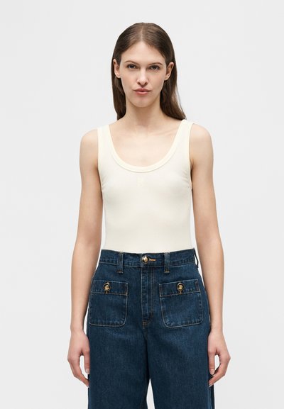 Femme aux longs cheveux bruns portant un haut blanc sans manches et un pantalon en denim bleu foncé taille haute avec des poches avant boutonnées.