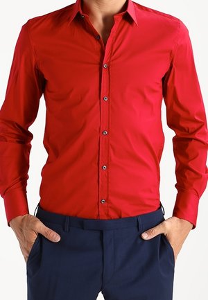 Mann trägt ein leuchtend rotes langärmeliges Hemd, das in dunkelblaue Hosen gesteckt ist, mit den Händen in den Taschen.