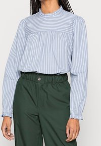 Blouse à rayures bleu clair avec encolure à volants et manches longues, associée à un pantalon vert taille haute avec une ceinture élastique.