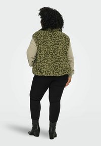 Person med krøllete hår iført en olivengrønn leopardmønstret vest, beige langermet skjorte, svarte bukser og svarte støvler med hæl, stående med ryggen til.