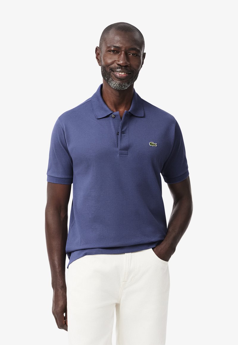 Polo blu navy in cotone con maniche corte, due bottoni, piccolo logo verde Lacoste, abbinato a pantaloni bianchi.
