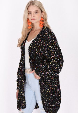 Zwarte gebreide cardigan met veelkleurige draadjes, open voorkant, lange mouwen en een gemêleerde stof. Gecoördineerd met een witte top en lichtblauwe jeans.