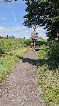 Bėgikas pilku beisbolo triuku ir juodomis šortais ant žvyrkelio, apsupto žalios žolės ir medžių, po aiškiai mėlynu dangumi.
