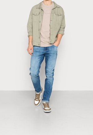 Lichtgroene spijkerjas over een beige T-shirt, gecombineerd met verweerde blauwe jeans en wit met bruine details sneakers. Casual, gelaagde outfit.