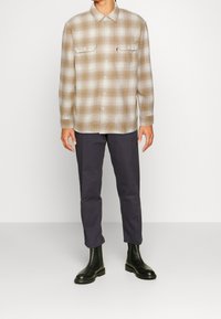 Camicia di flanella a quadri beige con maniche lunghe e due tasche con bottone, abbinata a pantaloni blu scuro e stivali neri alla caviglia.