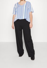 Henkilö, joka käyttää löysää sinivalkoista mallikuvioista lyhythihaista paitaa, mustia leveälahkeisia housuja ja mustia neliönmuotoisia kenkiä, seisoo harmaalla lattialla.