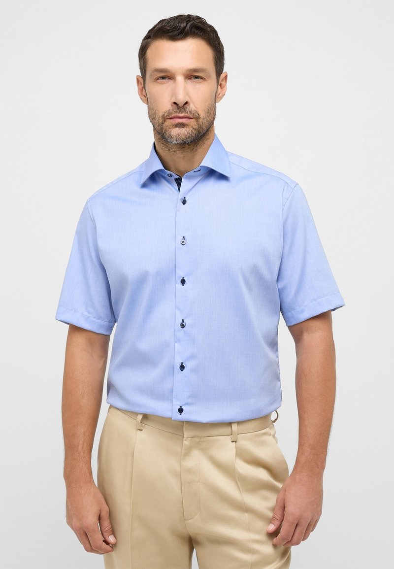 Lichtblauw kortärmshirt met een gestructureerde kraag, zwarte knopen en een soepele stoftextuur, gedragen met beige broeken.