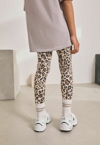 Leopárd mintás leggings bézs alapszínnel, rugalmas anyagból, fehér sportcipőkkel és csíkos bokazoknival párosítva, egy túlméretezett szürke pólóval kiegészítve.