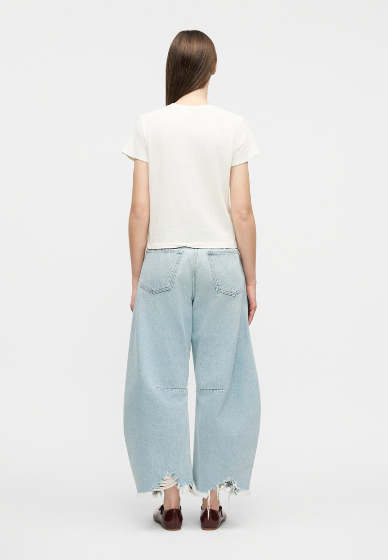 Frau mit glatten braunen Haaren, die ein weißes kurzärmeliges Oberteil und weit geschnittene, hellblaue Jeans mit Used-Optik trägt, stehend vor einem schlichten Hintergrund.