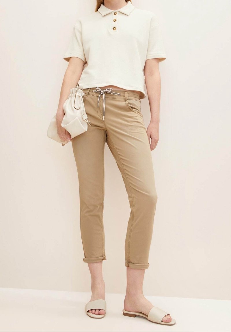 Polo-shirt crème coupé avec trois boutons marron, pantalon beige cintré avec cordon de serrage et sandales claires. Petit sac à main blanc comme accessoire.