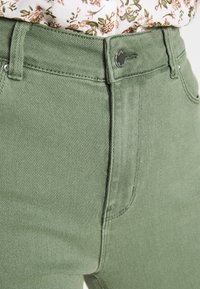 Pantalones de mezclilla verde con un acabado texturizado, cintura ajustada y detalle de botón metálico. Costuras visibles a lo largo de las costuras, combinados con una blusa floral.