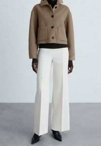 Veste courte en laine marron avec deux poches avant, boutons noirs et col de chemise, assortie à un pantalon large blanc et des bottes noires.