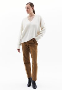 Maglione oversized a scollo a V crema abbinato a pantaloni marroni su misura e stivaletti neri con finitura testurizzata. Il modello è in piedi contro uno sfondo bianco.