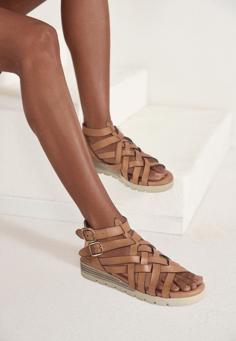 Personne portant des sandales en cuir tressé marron avec des brides à double boucle sur un sol blanc près de marches blanches.
