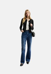 Gilet noir à boutons sur une chemise blanche, associé à un jean évasé bleu ; modèle tenant un petit sac à bandoulière noir. Chaussures noires.