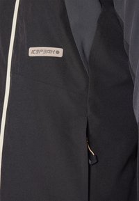 Czarna kurtka na zewnątrz z jasnoszarym zamkiem i naszywką z logo. Materiał ma gładką teksturę i elegancki fason.