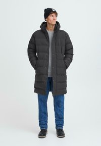 Svart puffjakke med hette, med et quiltet design og frontknapper, brukt over en grå genser og blå jeans.