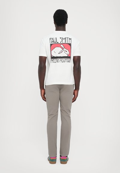 Vit t-shirt med kort ärm som visar en grafisk bild av en kanin och text på baksidan; kombinerad med ljusbeige byxor och flerfärgade sneakers.