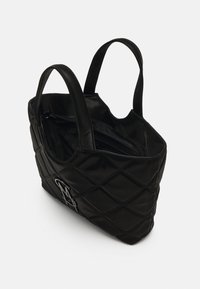 Sac fourre-tout noir matelassé avec doubles poignées et un emblème métallique sur le côté, présenté partiellement ouvert sur un fond blanc.