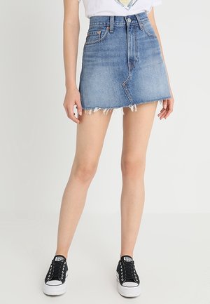 Mujer con una minifalda vaquera asimétrica azul con dobladillo deshilachado, camiseta blanca y zapatillas negras y blancas, de pie sobre un suelo blanco.