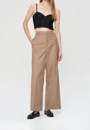 Femme portant un top court noir à fines bretelles, un pantalon beige taille haute à jambes larges et des mocassins noirs à enfiler avec des détails dorés.