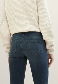 Vaqueros de mezclilla azul oscuro con un diseño ajustado, cuentan con dos bolsillos traseros y una textura suave, combinados con un suéter crema de textura.