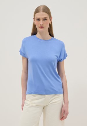 Vrouw met lang blond haar draagt een lichtblauw kortermig shirt en gebroken witte broek, staande tegen een effen lichte achtergrond.